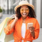 glad-excited-shocked-young-black-woman-casual-with-glasses-hat-with-open-mouth-hold-many-bags-phone