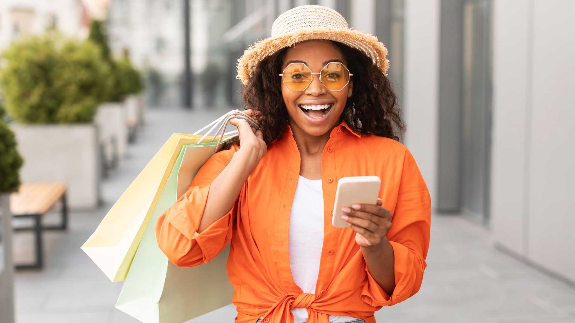 glad-excited-shocked-young-black-woman-casual-with-glasses-hat-with-open-mouth-hold-many-bags-phone
